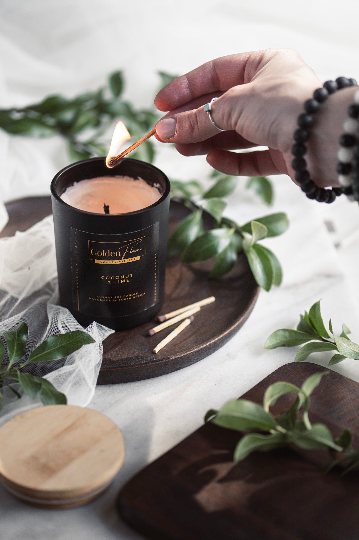 Hand lighting a candle with a match, surrounded by greenery on a light surface.